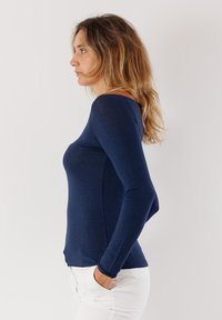 Top aderente a maniche lunghe blu navy realizzato in tessuto morbido, con scollatura a barca e vestibilità slim, abbinato a pantaloni bianchi e visto di profilo.