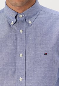 Camisa abotonada azul claro con un sutil patrón texturizado, botones blancos y un pequeño logotipo en la zona del pecho con detalles en rojo, blanco y azul.