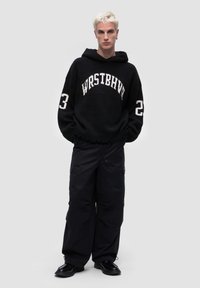 Mustat neulehuppari, jossa on "WRSTBHVR" -teksti ja numero 23. Yhdistetty mustiin cargo-housuihin. Malli seisoo vaaleanharmaata taustaa vasten.