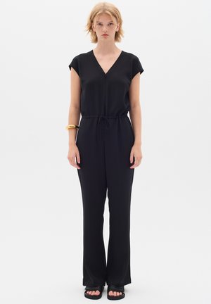 Combinaison noire à manches courtes, décolleté en V, taille à cordon et jambes larges. Tissu lisse, associée à des sandales noires et un bracelet en or.