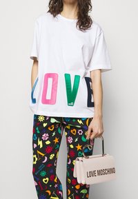 Vit bomull T-shirt med färgglad "LOVE" text, ihop med svarta byxor med livliga mönster. Håller en ljusrosa handväska med logotyp.