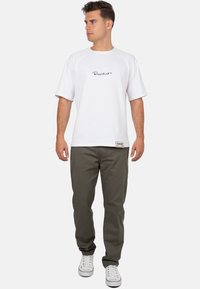 T-shirt blanc à manches courtes avec logo noir, associé à un pantalon vert olive et des baskets blanches. Le t-shirt a un col rond et une coupe décontractée.
