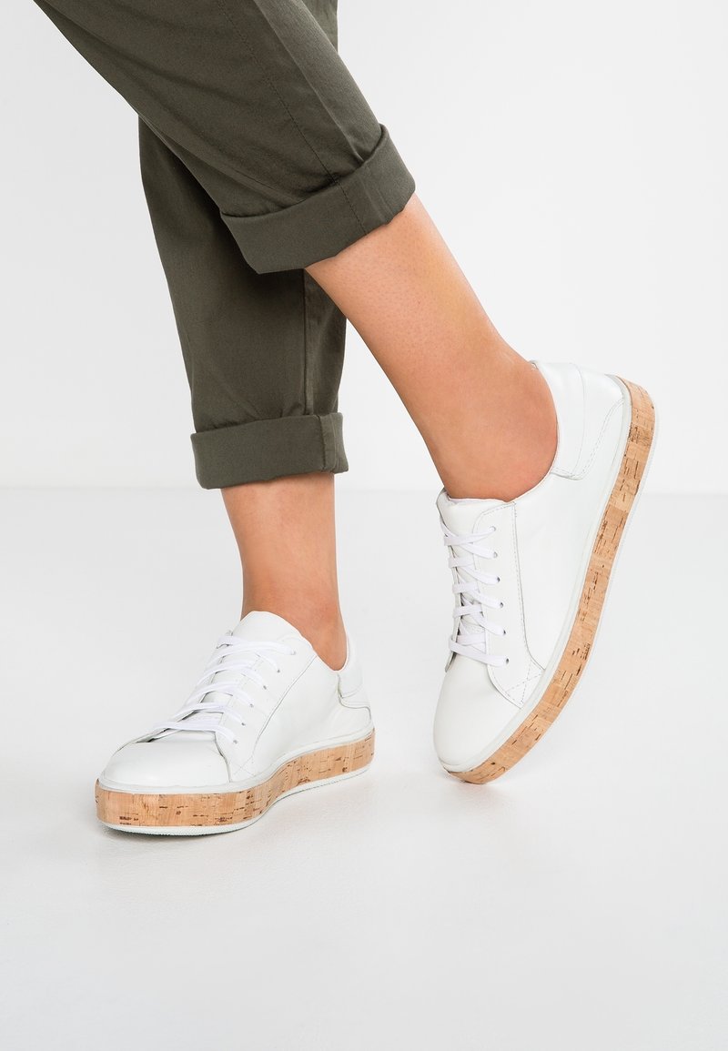 Vita lädersneakers med en texturerad korkplattform. Snörning och minimal sömdetalj, tillsammans med rullade olivgröna byxor med uppvikta ben.