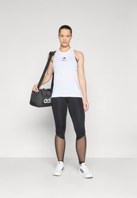 Top de sport alb cu logo negru, asortat cu colanți negri cu panouri din plasă și pantofi albi. Este inclus o geantă de sport neagră.