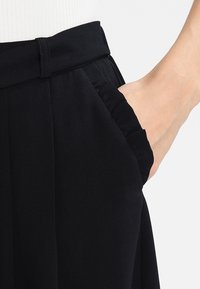 Pantalons noirs taille haute en tissu texturé, avec une poche latérale et un détail de taille froncée.