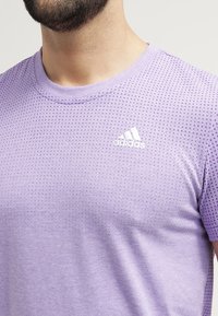 Fialové sportovní tričko vyrobené z lehkého materiálu, se strukturovaným vzorem a bílým logem Adidas na hrudi.