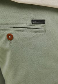Heledamad rohelised püksid on valmistatud tekstuurilisest kangast, millel on väike must silt "JACK & JONES" ja pruuni nupuga aktsent tagumise tasku lähedal.