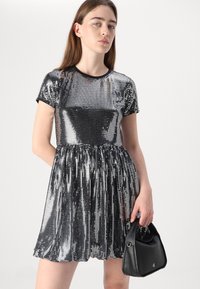 Sidabrinė sequin suknelė su prigludusia liemene ir platėjančia sijoną. Ją lydi maža juoda rankinė su vingiuotu dizainu ir subtiliu logotipu.