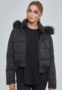 Svart kort puffjacka med huva med fuskpäls, quiltat mönster och ribbad nederkant. Bärs över en svart skjorta med rund halsringning.