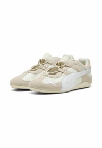 Zapatillas atléticas beige hechas de ante y malla, con acentos blancos. El diseño incluye un perfil bajo con cordones elásticos y una suela texturizada.
