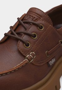 Καφέ δερμάτινο παπούτσι τύπου boat με στρογγυλά καφέ κορδόνια, μεταλλικές οπές, λεπτομερή ραφή και ταμπέλα με λογότυπο στο πλάι σε λαστιχένια σόλα.