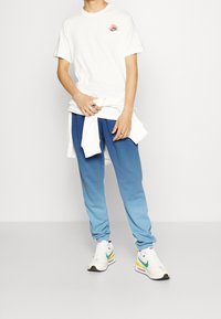 Bílý krátký rukáv tričko s růžovým a černým logem, spárováno s modrými gradientovými tepláky. Bílé tenisky s zelenými a žlutými akcenty.