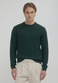 Grüner strukturierter Strickpullover mit rundem Ausschnitt und gerippten Bündchen, kombiniert mit cremefarbenen, plissierten Hosen.