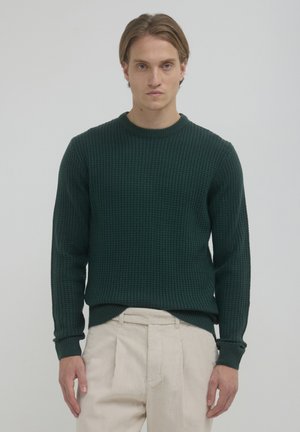 Grüner strukturierter Strickpullover mit rundem Ausschnitt und gerippten Bündchen, kombiniert mit cremefarbenen, plissierten Hosen.