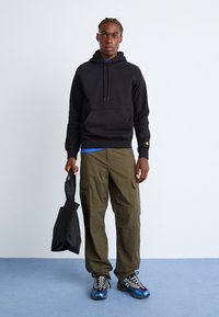 Un sweat à capuche noir avec une poche kangourou, un pantalon cargo olive avec des poches latérales et un sac noir. Portant des baskets grises et bleues avec un design à motifs.