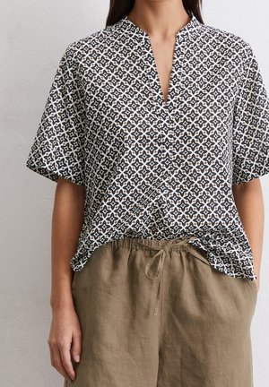 Top à manches courtes avec un décolleté en V, présentant un motif géométrique noir et blanc sur un tissu léger ; associé à un pantalon léger beige.