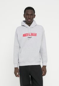 Γκρι φούτερ με κουκούλα και τσέπη καγκουρό, με κόκκινο γράφημα "METAL GEAR" στον μπροστινό μέρος. Κατασκευασμένο από μαλακό ύφασμα, με ριμπ μανσέτες.