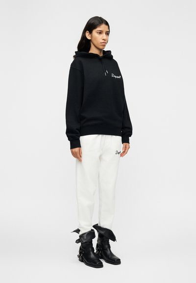 Sweat-shirt à capuche noir avec logo brodé, associé à un pantalon de jogging blanc avec broderie similaire. Bottines noires à boucles.