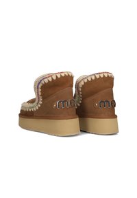 Botas de ante marrón hasta el tobillo con forro de piel de oveja, costuras decorativas y suela elevada. Presentan acentos de logotipo metálico y detalles de cuentas coloridas.