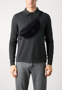 Svart kordfløyels fanny pack båret over brystet, med glidelås og logodetalj, kombinert med en mørkegrå langermet skjorte.