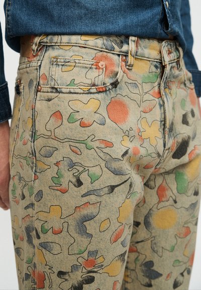 Gros plan sur un jean beige délavé avec des motifs floraux et feuillus abstraits colorés en rouge, jaune, vert et noir, porté avec une chemise en jean bleue.
