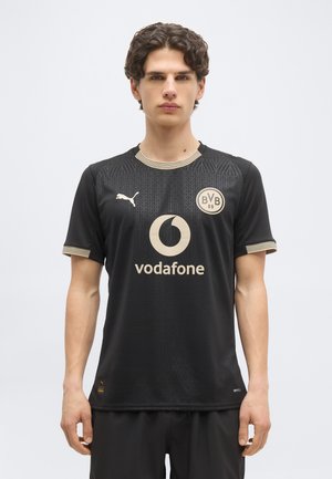 Joven con una camiseta negra del Borussia Dortmund de fútbol, con el logo beige de Vodafone y el emblema de Puma, posando frente a un fondo liso y claro.
