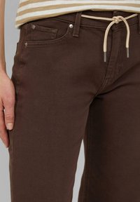 Brązowe dżinsy mają elastyczny pas z metalowymi akcentami oraz kieszenie z przodu. Materiał wydaje się być trwały i ma gładką teksturę.