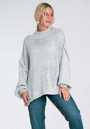Jasnoszary sweter z dzianiny o wysokim kołnierzu, luźnym kroju i opadających rękawach. Teksturowany wzór, surowe wykończenie i prążkowane mankiety. W połączeniu z niebieskimi dżinsami.