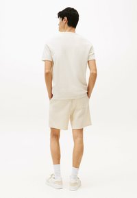 T-shirt beige chiaro a maniche corte, abbinato a pantaloni corti beige chiaro con texture. Sneakers bianche e calzini completano l'outfit; design minimalista.
