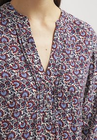 Blouse à imprimé floral avec des motifs bleus, rouges et blancs, dotée d'un col en V, de boutons sur le devant et d'un tissu texturé.