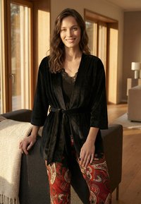 Glimlachende vrouw in een zwarte fluwelen badjas en rood patroonbroek staat binnen, leunend op een grijze bank met een houten vloer en grote ramen op de achtergrond.