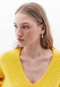 Geel, geribbeld sweater met een V-hals ontwerp. Het model heeft zilveren druppelvormige oorbellen. Het haar is steil en valt over de schouders met een lichte textuur.
