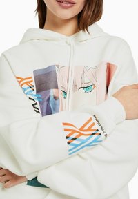 Vit hoodie med en grafisk bild av en karaktär med blå ögon, accentuerad med färgglada mönster på ärmarna i orange, svart och blå.