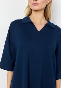 Jūras spēku zils polo stila tops ar V-veida kaklu, īsām piedurknēm un brīvu piegriezumu. Gluda tekstūra, minimālisma dizains un apkakles akcents.