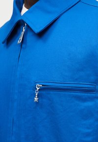 Κοντινό πλάνο για το γιακά μπλε πουκάμισου με μεταλλικό φερμουάρ που έχει ένα αστέρι ως έλκτρα στην τσέπη στο στήθος και στο κύριο φερμουάρ μπροστά.