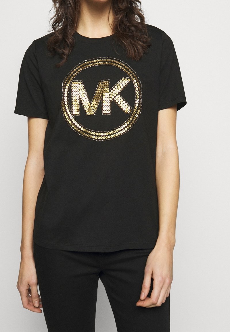 Svart t-shirt med korta ärmar som har en stor guld- och silverpaljetterad "MK"-logotyp i ett cirkulärt mönster på framsidan.