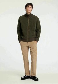 Ελιά πράσινη ζακέτα με φερμουάρ και ψηλό γιακά, υφασμάτινη επιφάνεια και ριχτές μανσέτες, συνδυασμένη με μπεζ παντελόνι και μαύρα παπούτσια.