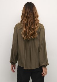 Blouse vert olive avec des manches longues en ballon et des détails en dentelle sur le plastron, présentant une coupe décontractée et une texture douce.