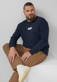 Marineblaues langärmliges Hemd mit grafischem Aufnäher, kombiniert mit braunen Hosen und weißen Sneakern. Die Textur wirkt glatt und hat einen lässigen Schnitt.