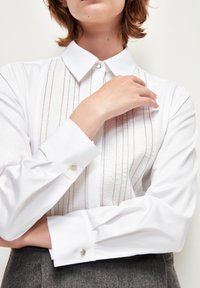 Camisa blanca con cuello estructurado, con rayas verticales texturizadas y acentos de botones plateados en los puños. Tejido suave.