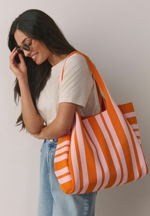 Femeie cu păr lung și închis la culoare, purtând ochelari de soare, cămașă bej, blugi albaștri, zâmbitoare și ținând o geantă mare de umăr în dungi portocalii și roz.