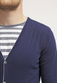 Marinblå cardigan med V-ringning, med knäppning fram och kontrasterande ljus trim. Bärs över en grå och vit randig skjorta.