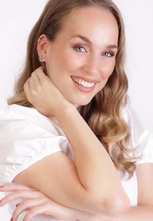 Nahaufnahme einer Person mit langen, welligen Haaren, die eine weiße Bluse trägt. Ihre Ohrringe haben ein silbernes, geschwungenes Design mit kleinen Verzierungen.
