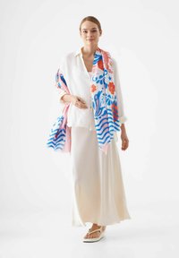 Écharpe légère et colorée avec des motifs floraux et de vagues, drapée sur une chemise en lin blanche et une jupe fluide crème, accompagnée de sandales.
