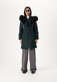Padded green parka with black faux fur trim on the hood, quilted texture, front zipper, and side pockets. Worn with gray wide-leg pants.