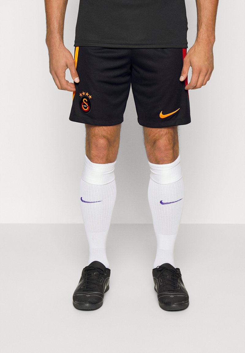 Shorts de football noirs avec des accents orange et rouge, ornés d'un logo et d'étoiles. Associés à des chaussettes blanches et des chaussures de sport noires.