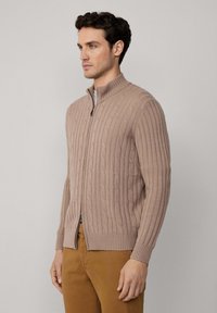 Beżowy kardigan z dzianiny w warkocze z wysokim kołnierzem, zamkiem błyskawicznym, ściągaczami u dołu i na rękawach, z teksturowanymi wzorami w całej konstrukcji.