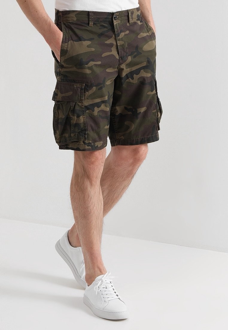 Man in groene en bruine camouflage cargo shorts en witte sneakers, staande met zijn handen in de zakken tegen een effen achtergrond.