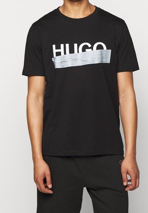 Czarna koszulka z krótkim rękawem z białym napisem "HUGO" częściowo zakrytą srebrną taśmą na klatce piersiowej, noszona z czarnymi spodniami.