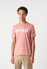 Rožinės spalvos medvilnės marškinėliai trumpomis rankovėmis su dideliu baltu "HUGO" logo priekyje, apvaliu iškirpimu ir laisvu siluetu.